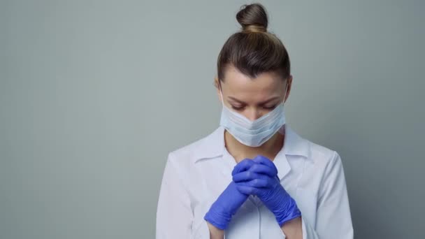A female doctor therapist in a white robe, mask and gloves. Face close-up. The doctor cries and prays. Tears in eyes. Pandemic and virus epidemic. Coronavirus covid-19. - Кадры, видео
