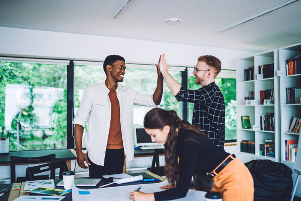 Side view of young woman at table with smiling multiethnic men giving high five in modern office being happy with successful work - Foto, immagini