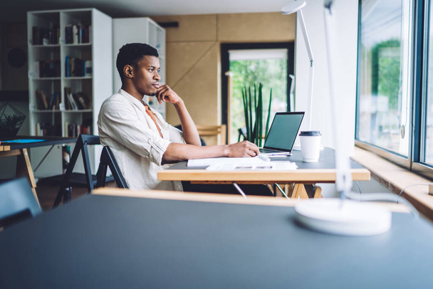 Young African American man sitting at table with laptop in office and looking thoughtfully away in window while dreaming in daylight - Foto, Bild