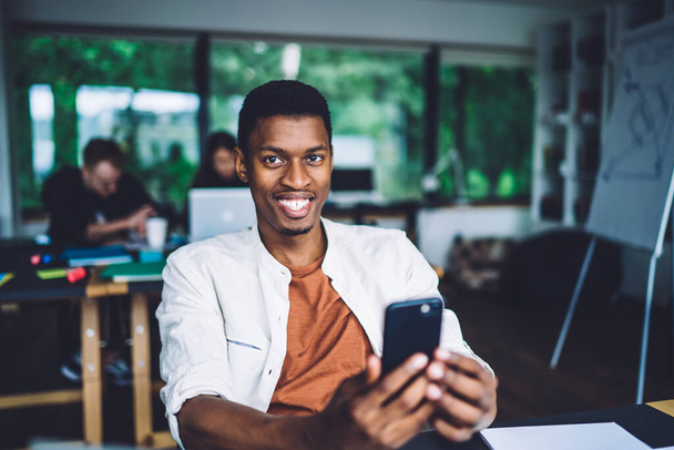 Young casual African American man smiling away while sitting at working desk in office with colleagues on backdrop and holding smartphone - Valokuva, kuva