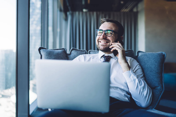 Happy male director in eyewear for vision correction feeling good from received good news from secretary during phone conversation indoors, cheerful man smiling while talking with colleague via app - Zdjęcie, obraz