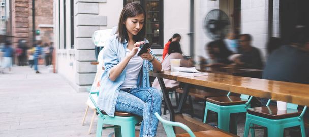 Attractive Chinese woman in casual denim outfit checking delivery information on literature web store while sitting at cafe terrace with public wifi internet, millennial hipster girl blogging - Zdjęcie, obraz