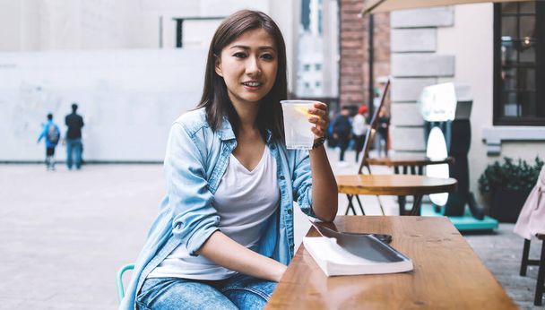 Portrait of positive Chinese female in casual denim shirt holding glass with cold lemon water and looking at camera during leisure day in downtown, attractive woman with beverage in hand outdoors - Photo, image