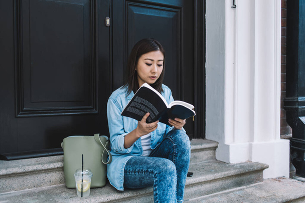 Young beautiful female tourist reading interesting book during rest after walking in city downtown, charming woman student preparing to study at university while is sitting near house door - Foto, Imagem