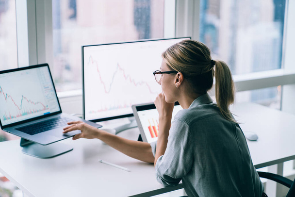 Side view of contemporary blond woman in glasses sitting at desktop in modern office using laptop and watching statistical graphs  - Фото, изображение