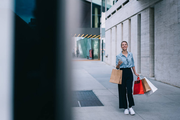 Positive young woman dressed in casual wear walking outdoors and doing shopping in black friday enjoying big sales and leisure time, hipster girl with paper bags and cellular strolling on street - Fotografie, Obrázek