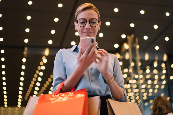 Cheerful female shopaholic holding shopping bags and looking at smartphone screen during online chatting after day in mall, successful hipster girl in optical spectacles for vision correction - Foto, Bild