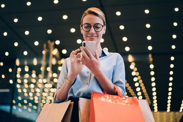Happy millennial female shopaholic continue shopping online via smartphone gadget after time in mall, young positive hipster girl with bags reading news about Black Friday via cellular phone - Фото, зображення
