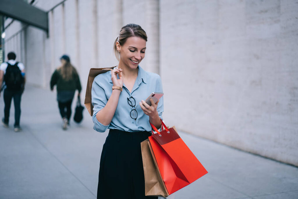 Happy woman using smartphone for sending text messages chatting via modern application, female customer with packages sharing publication about Black Friday sales in personal fashion web blog - Fotó, kép