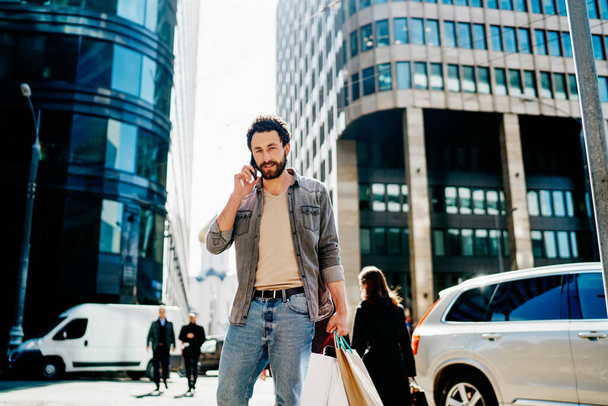Half length portrait of handsome hipster guy in trendy wear having mobile phone conversation standing on street with shopping bags, bearded man calling via cellphone after buying purchase in cit - Valokuva, kuva