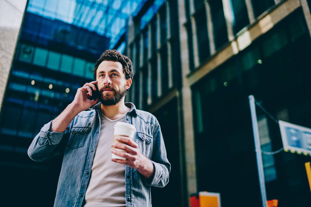 Bearded young man dressed in casual wear holding tasty coffee to in hand while calling on smartphone and walking in business center.Hipster guy communicating on mobile phone device on street - Foto, afbeelding