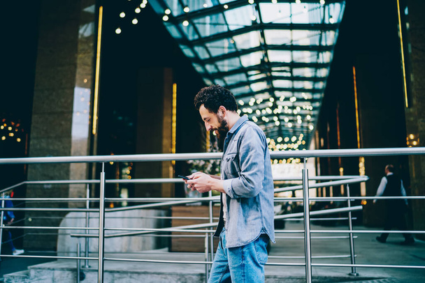 Serious caucasian male in casual wear walking with smartphone outdoors on urban settings, concentrated millennial man read notification on mobile phone connected to 4G strolling in downtow - Foto, immagini