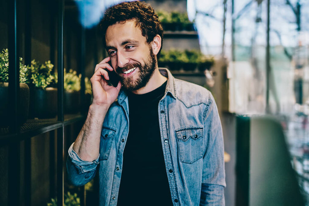Cheerful hipster student dressed in stylish denim casual wear laughing during phone conversation on smartphone.Positive bearded young man talking with friend on cellular standing in coworking space - Foto, immagini