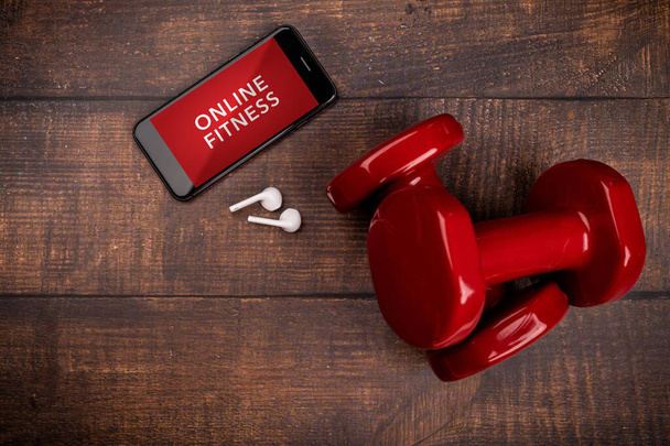 Piros súlyzó és okostelefon fából készült háttérrel. Alkalmazás a belső edzésre. Online Fitness program. Otthon, online edzés. Felülnézet, Covid-19 Coronavirus karantén koncepció - Fotó, kép