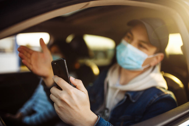 Мужчина в медицинской маске держит мобильный телефон в руке во время вождения автомобиля. Водитель проверяет новости о коронавирусе в пробке. Изоляция и здравоохранение во время карантинной концепции
 - Фото, изображение