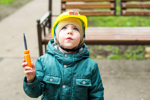Cheerful boy is standing on the street looking up, yellow helmet is on his head, a screwdriver is in his hand. Child plays a builder. Concept of protection from external influences - Фото, зображення