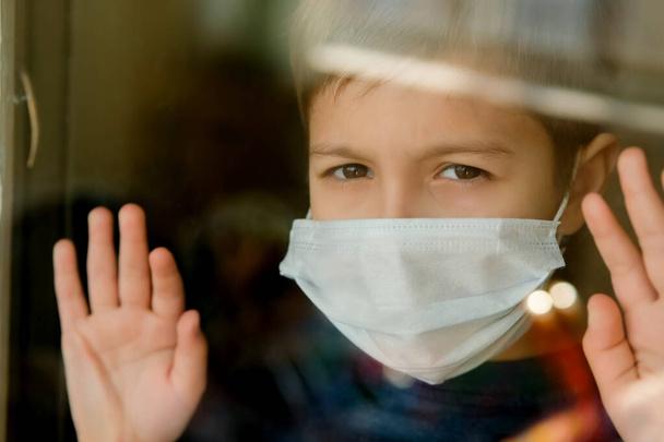 Young boy in a medical mask looks out the window. Self-isolation in quarantine, coronavirus, covid 19. - Foto, imagen