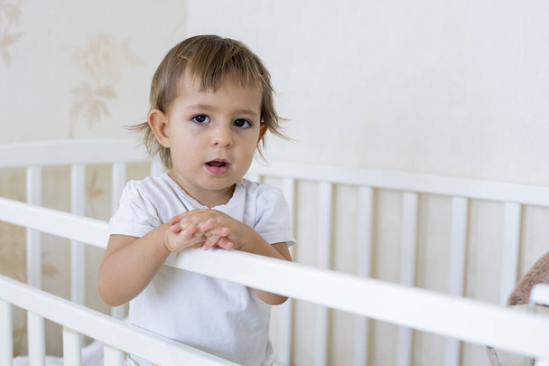concept covid-19, stay home quarantined, take care of your family. little cute toddler is standing in the crib at home and looking at the camera - Zdjęcie, obraz