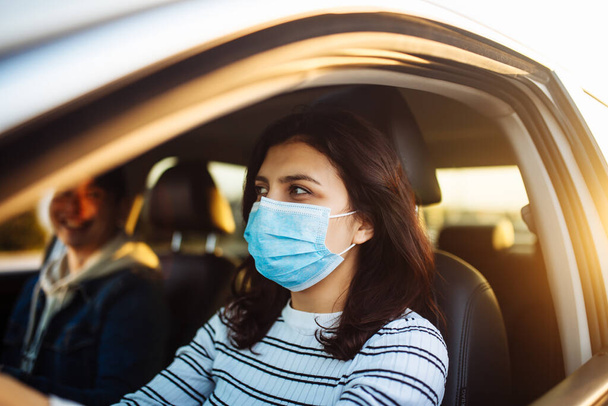 A girl drives a car with a passanger wearing medical mask during coronavirus pandemic quarantine. Woman taxi driver stuck in a traffic jam. Stay safe and healthcare concept - Foto, afbeelding