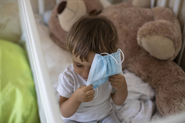 koncepce karantény a sebeizolace v pandemii coronavirus covid -19. malé roztomilé dítě objímá lékařskou masku, zatímco sedí v kolébce doma na pozadí medvídka - Fotografie, Obrázek
