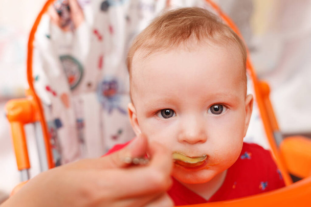 a small child eats with a spoon and sits and holds the edge of the feeding table in the nursery, grimy and stained with food, baby food concept, complementary foods, the first spoon - Zdjęcie, obraz