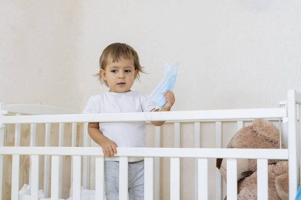 Пандемия коронавируса-ковида-19 карантина. Маленький милый серьезный ребенок в детской кроватке дома в безопасности и держит медицинскую маску. копировальное пространство
 - Фото, изображение