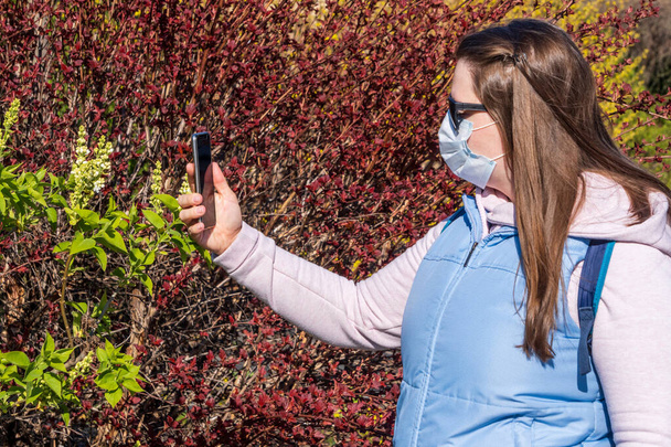 A girl in a medical mask and dark glasses takes a selfie or broadcasts live on the Internet, against background of spring flowering bushes during a coronavirus pandemic or pollen Allergy. - Photo, Image