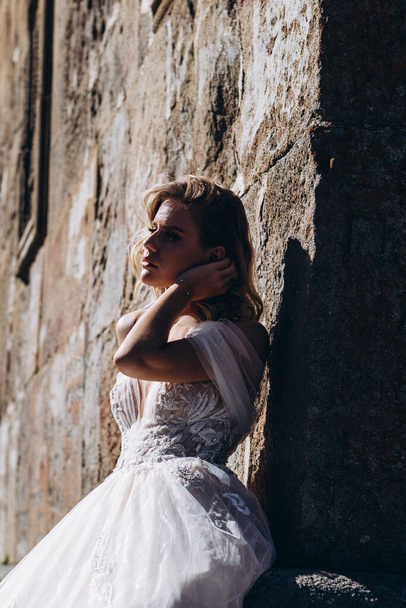 young bride dressed in a long white dress posing for a photo on a background of a large stone wall, a stylish photo for the promotion of women's dress salon - Zdjęcie, obraz