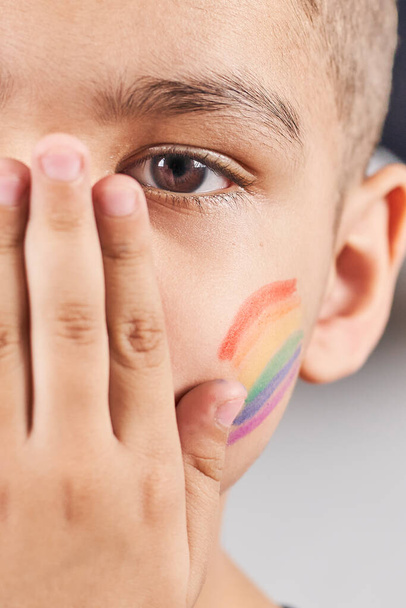 頬に虹の旗を描いた小さな男の子の半分の顔のクローズアップビュー。LGBTQファミリーの子供の顔の肖像画。プライドの日、 LGBT家族、性的権利と平等の概念 - 写真・画像