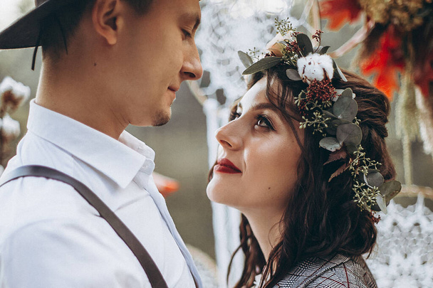 01.06.2019 Tbilisi, Georgia: stylish couple of brides: man in tuxedo and hat, woman in light white dress with wreath on head against evening park background, lovers posing at wedding photo shoot. wedding background - Fotó, kép