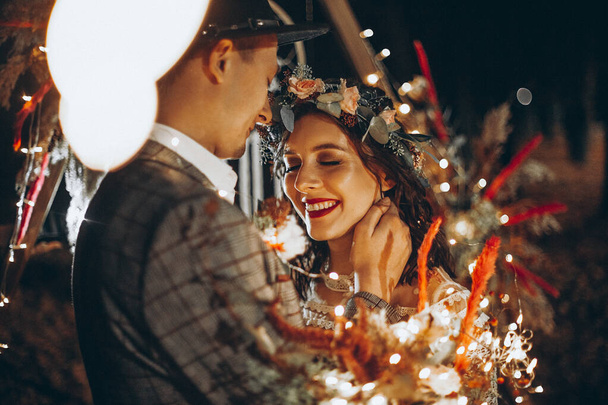 Portrait of a couple of young brides in front of a wedding arch at night. Romantic wedding ceremony in nature. The lights of the electric garland illuminate the wedding party. - Foto, Imagem