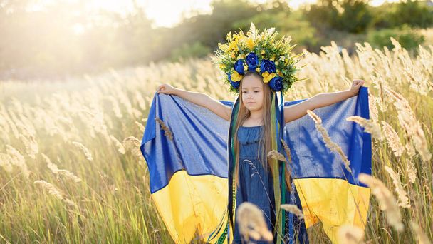 cute little girl wearing floral wreath with ribbons holding blue and yellow Ukrainian flag and walking at summer meadow, Ukraine Independence Day concept, Day of National Flag of Ukraine and Constitution day, Patriotic holiday   - Foto, imagen