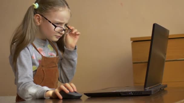 Linda niña en eyeglassess está sentada frente a su portátil negro y estudiar en casa debido a la pandemia de Coronavirus Covid-19 y auto-aislamiento. Concepto de educación remota
 - Metraje, vídeo