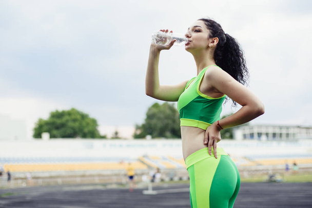Morena de pelo rizado practicando deportes en el estadio. Chica bebe agua de una botella después del entrenamiento
 - Foto, imagen