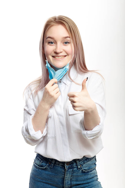 jong blond meisje in een witte blouse en neemt af een medisch masker op een witte achtergrond glimlachen en tonen duim omhoog - Foto, afbeelding