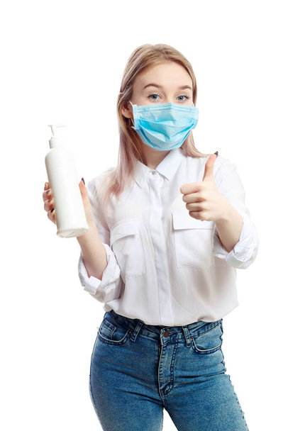 young blonde girl in a white blouse and a medical mask uses an antiseptic on a white background. - Foto, afbeelding