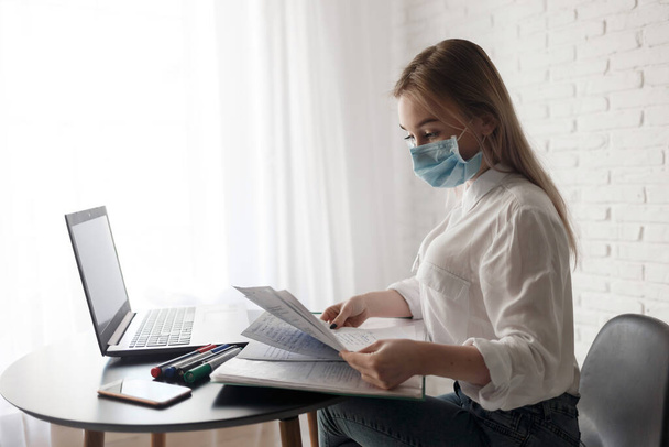 junges blondes Mädchen in weißer Bluse und medizinischer Maske arbeitet im Raum mit Dokumenten und Laptop. Arbeit zu Hause. Eine Frau sitzt an einem Tisch und hält Papier in den Händen - Foto, Bild