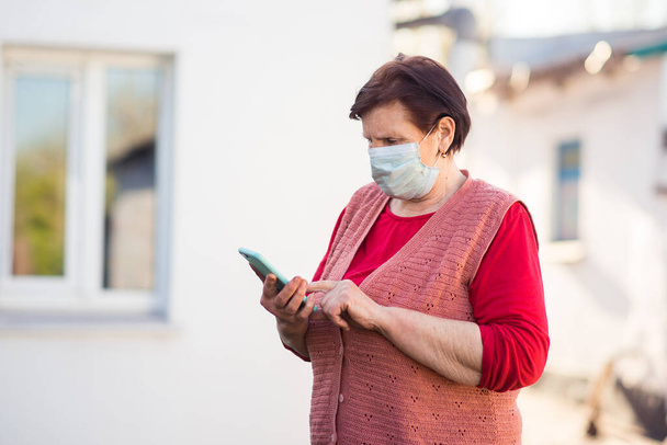 Woman in orange sweater and medical mask using her phone on empty street. Coronavirus pandemic concept. - Foto, afbeelding