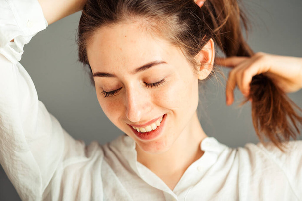 Een lachend meisje met een tevreden gezicht houdt haar haar met haar handen op een grijze achtergrond, close-up. Naakt make-up, zonder retoucheren. - Foto, afbeelding
