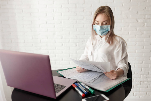 junges blondes Mädchen in weißer Bluse und medizinischer Maske arbeitet im Raum mit Dokumenten und Laptop. Arbeit zu Hause. Eine Frau sitzt an einem Tisch und hält Papier in den Händen - Foto, Bild