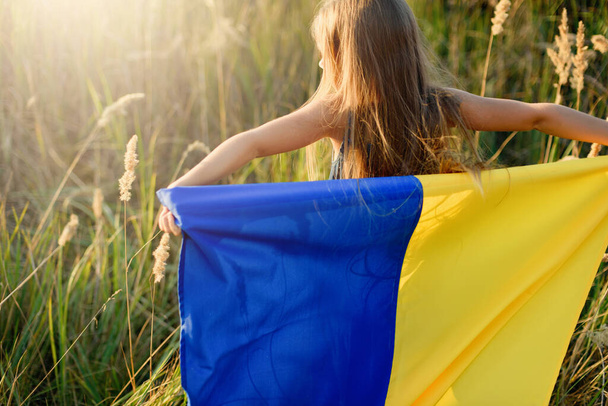 happy little girl with fluttering blue and yellow flag of Ukraine walking in field at sunny summer day, concept of Ukraine Independence Day and Constitution day, Patriotic holiday  - Foto, Bild