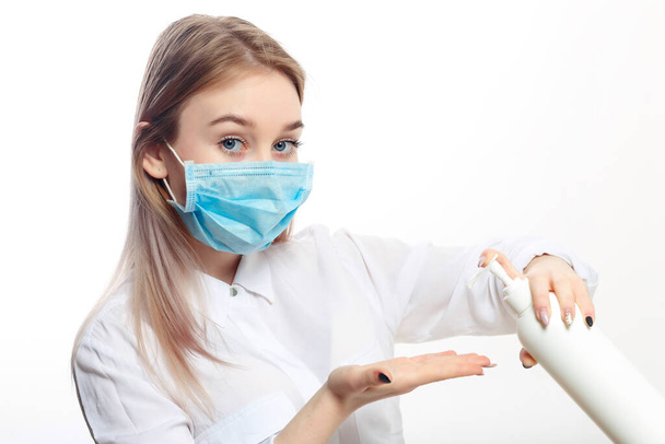 young blonde girl in a white blouse and a medical mask uses an antiseptic on a white background. - Foto, Imagem