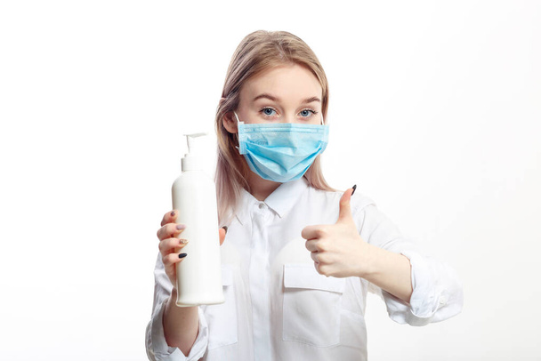 giovane ragazza bionda in una camicetta bianca e una maschera medica utilizza un antisettico su uno sfondo bianco
. - Foto, immagini