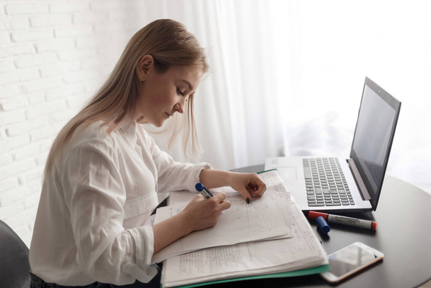 Молодая блондинка в белой блузке работает в комнате с документами и ноутбуком. Работать дома. Женщина смотрит в камеру, сидя за столом с документами
. - Фото, изображение