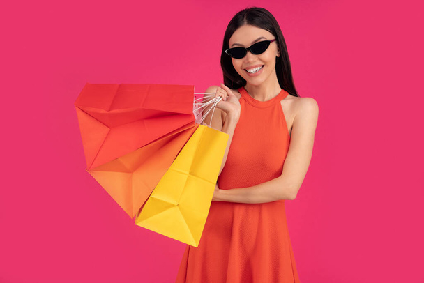 Immagine di una bella donna asiatica felice eccitata in posa isolata su sfondo rosa parete che tiene borse della spesa. vestito in abito arancione e occhiali da sole
. - Foto, immagini