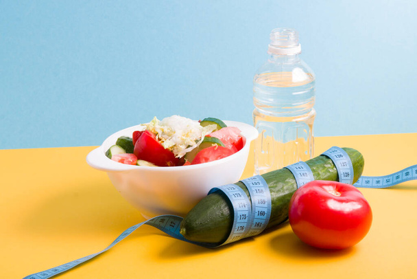 small bottle of water, salad in a white bowl, blue measuring tape around a cucumber, tomato on a blue and yellow background copy space, diet and weight loss concept - Foto, afbeelding