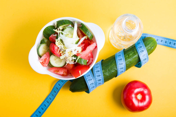 small bottle of water, salad in a white bowl, blue measuring tape around a cucumber, tomato on a blue and yellow background copy space, diet and weight loss concept, top view - Photo, Image