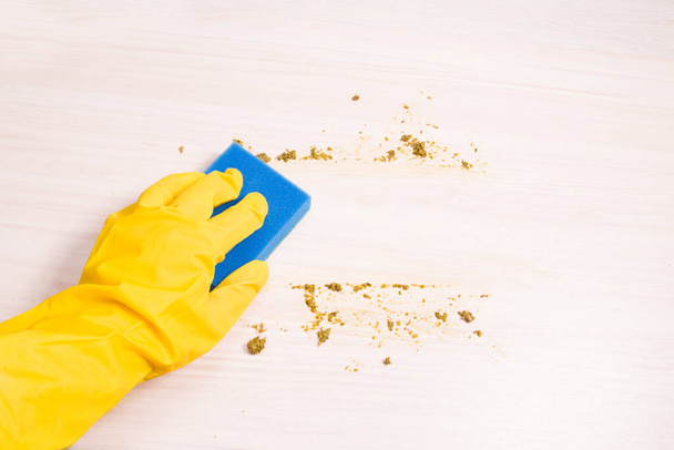 a female hand in a yellow rubber glove wipes with a blue sponge for washing dishes leftover food on a wooden table, cleaning after cooking, copy space, light background - Fotoğraf, Görsel