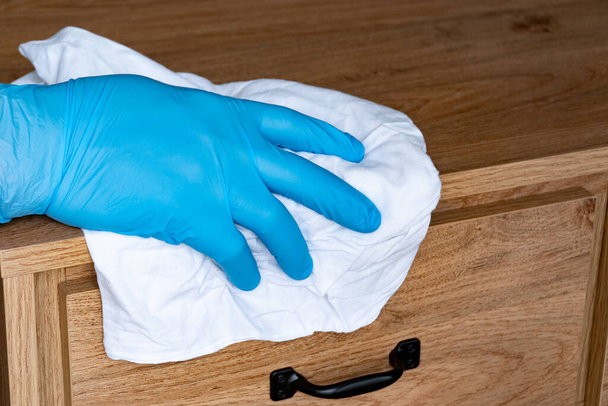 Zabraňte šíření koronaviru a infekce COVID-19. Muž v ochranných modrých rukavicích umývá nábytek na nočním stolku hadrem. - Fotografie, Obrázek