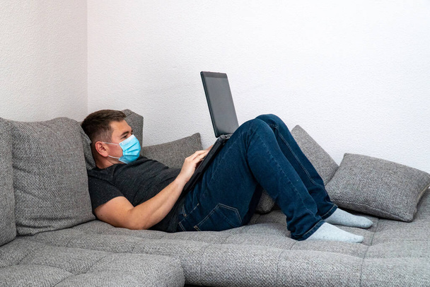 Ένας άνθρωπος σε καραντίνα με ιατρική μάσκα είναι κουρασμένος και ξαπλώνει σε έναν καναπέ για να δουλέψει στο λάπτοπ. Καραντίνα αυτοαπομόνωσης σε πανδημία στο σπίτι COVID19. - Φωτογραφία, εικόνα